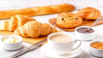Une table mise pour le petit-déjeuner avec des croissants, une baguette, du beurre, de la confiture, du café et un couteau.