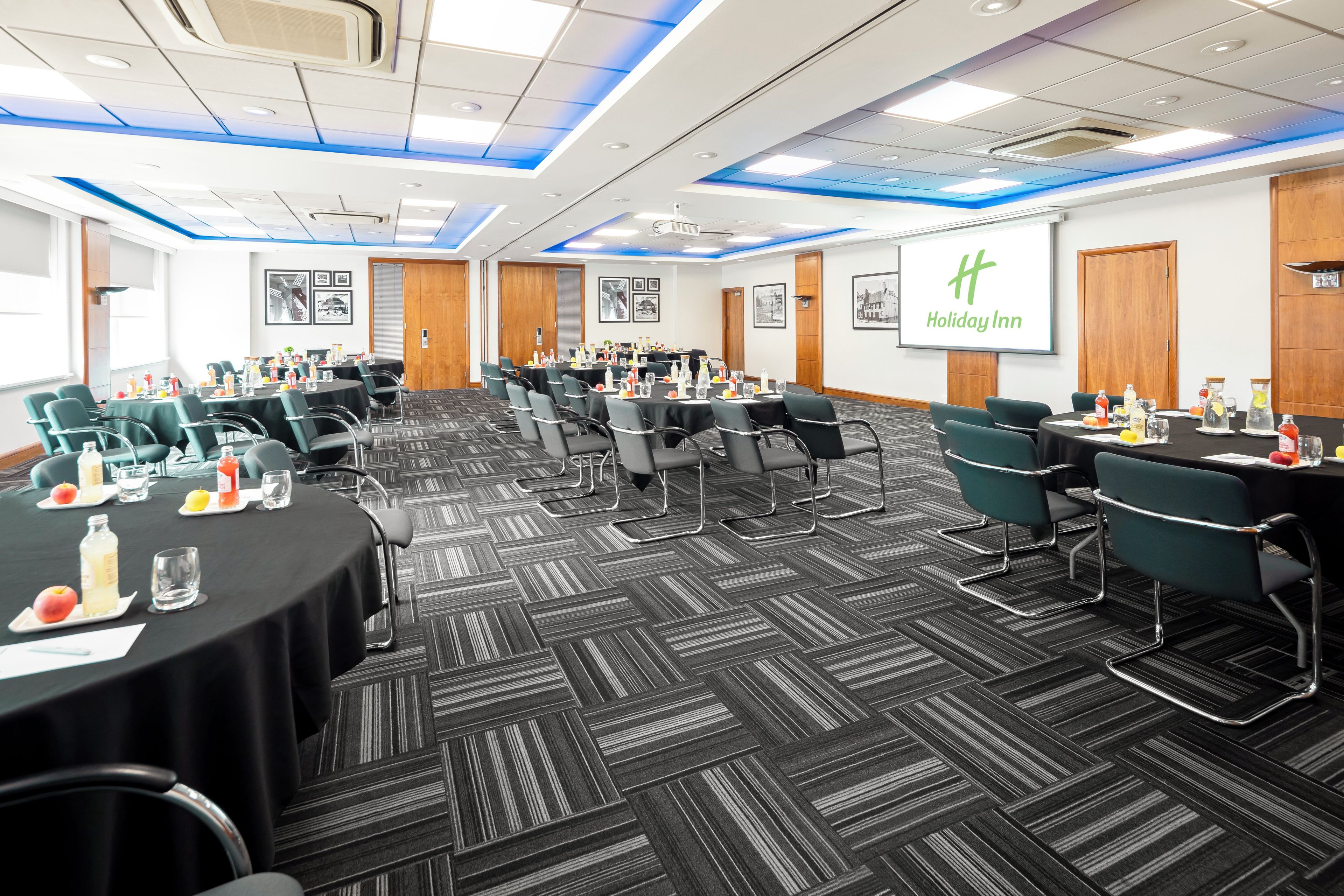 A conference room with multiple tables, chairs, and a large screen displaying the Holiday Inn logo.