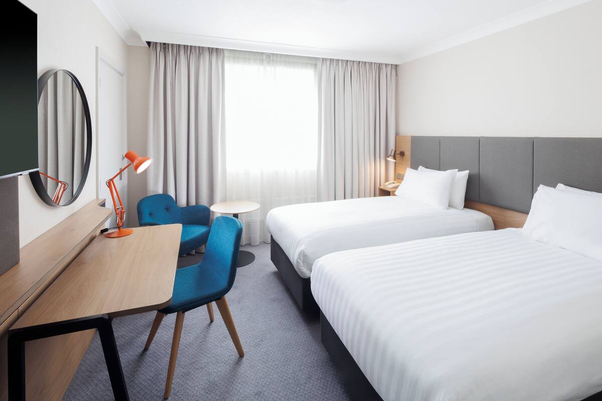 A hotel room with two beds, a desk, a chair, a lamp, and a large window with curtains.