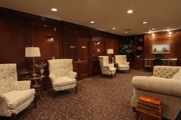 A hotel room with a large wooden wardrobe, four upholstered armchairs, a sofa, a small table with a lamp, and a bookshelf with books and a framed picture.