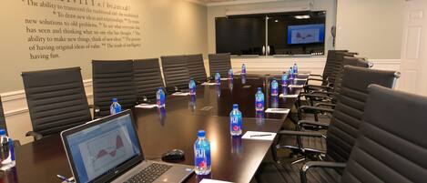 A conference room with a long table, black chairs, laptops, water bottles, and a screen displaying a graph.