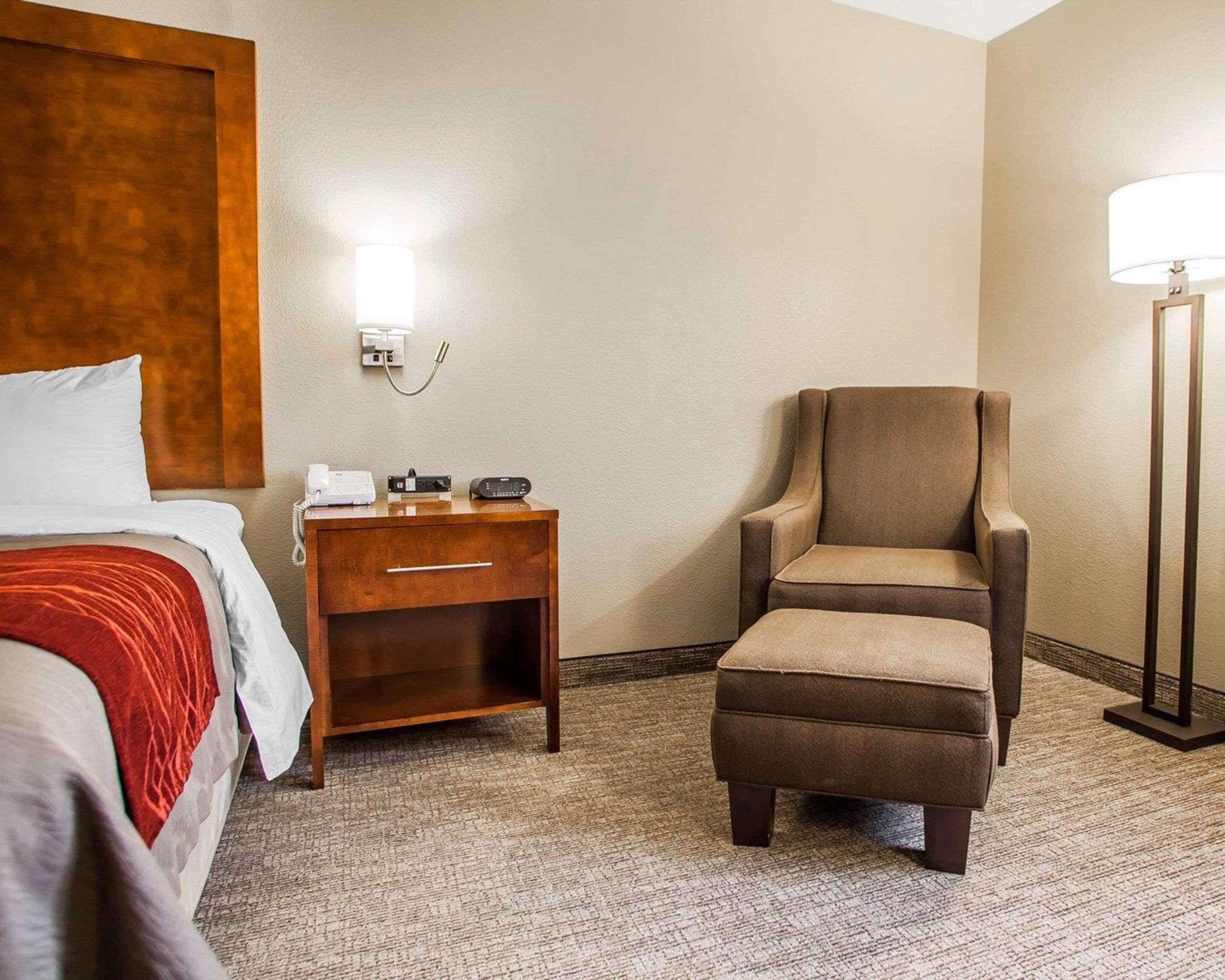 Een hotelkamer met een bed, nachtkastje met lamp, een stoel en een vloerbedekking.