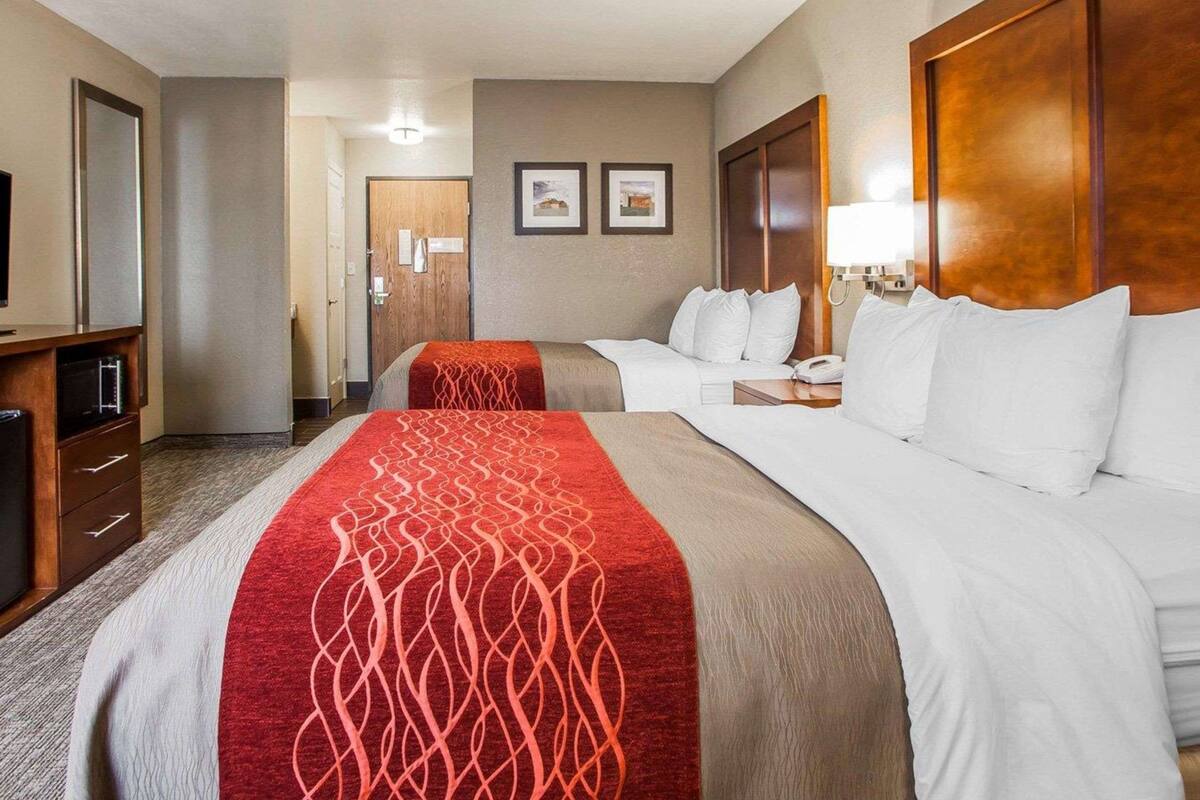 A hotel room with two beds, a desk, a TV, and a door leading to a hallway.