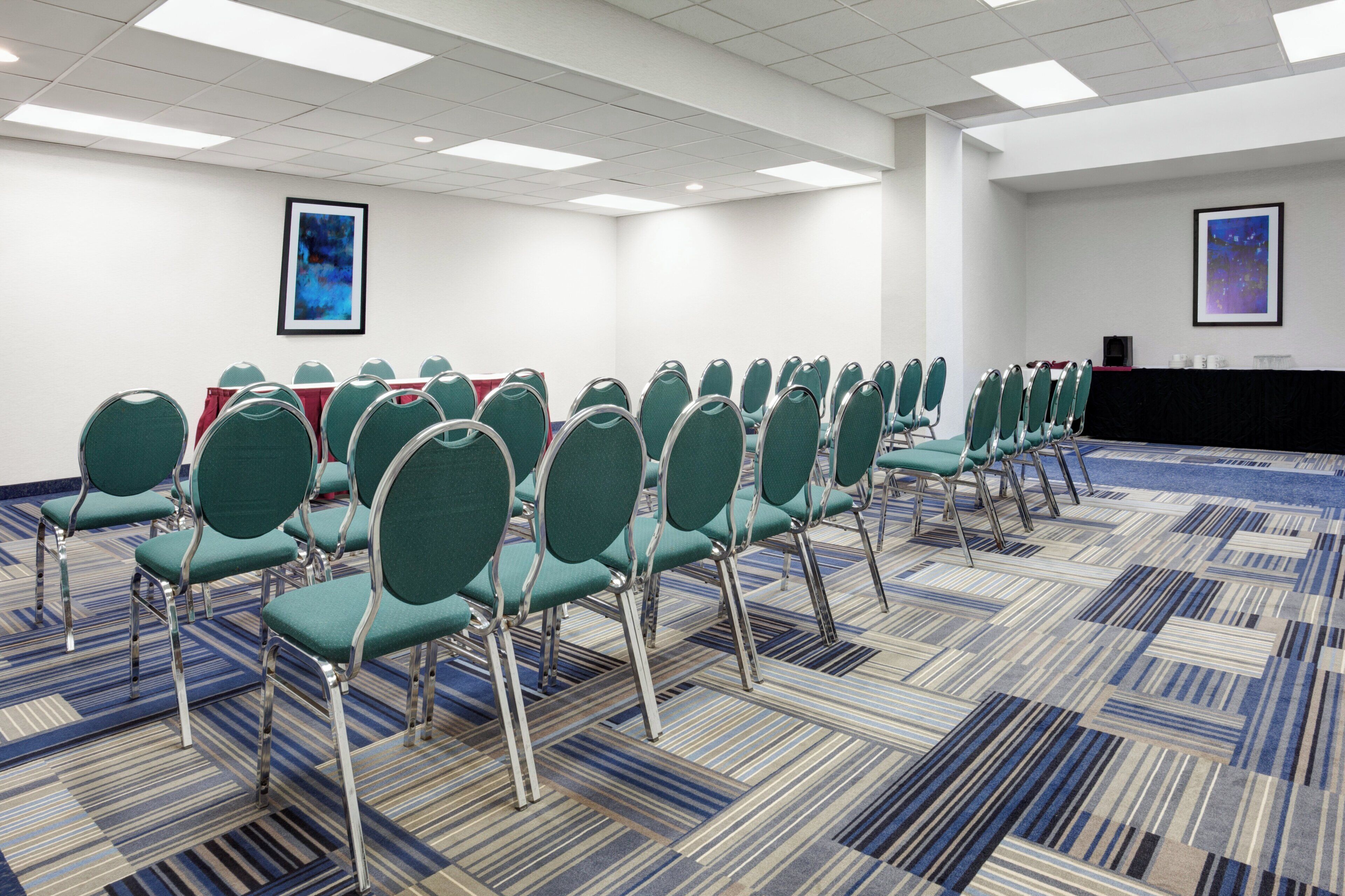 A conference room with rows of green chairs, a patterned carpet, and two framed abstract paintings on the walls.