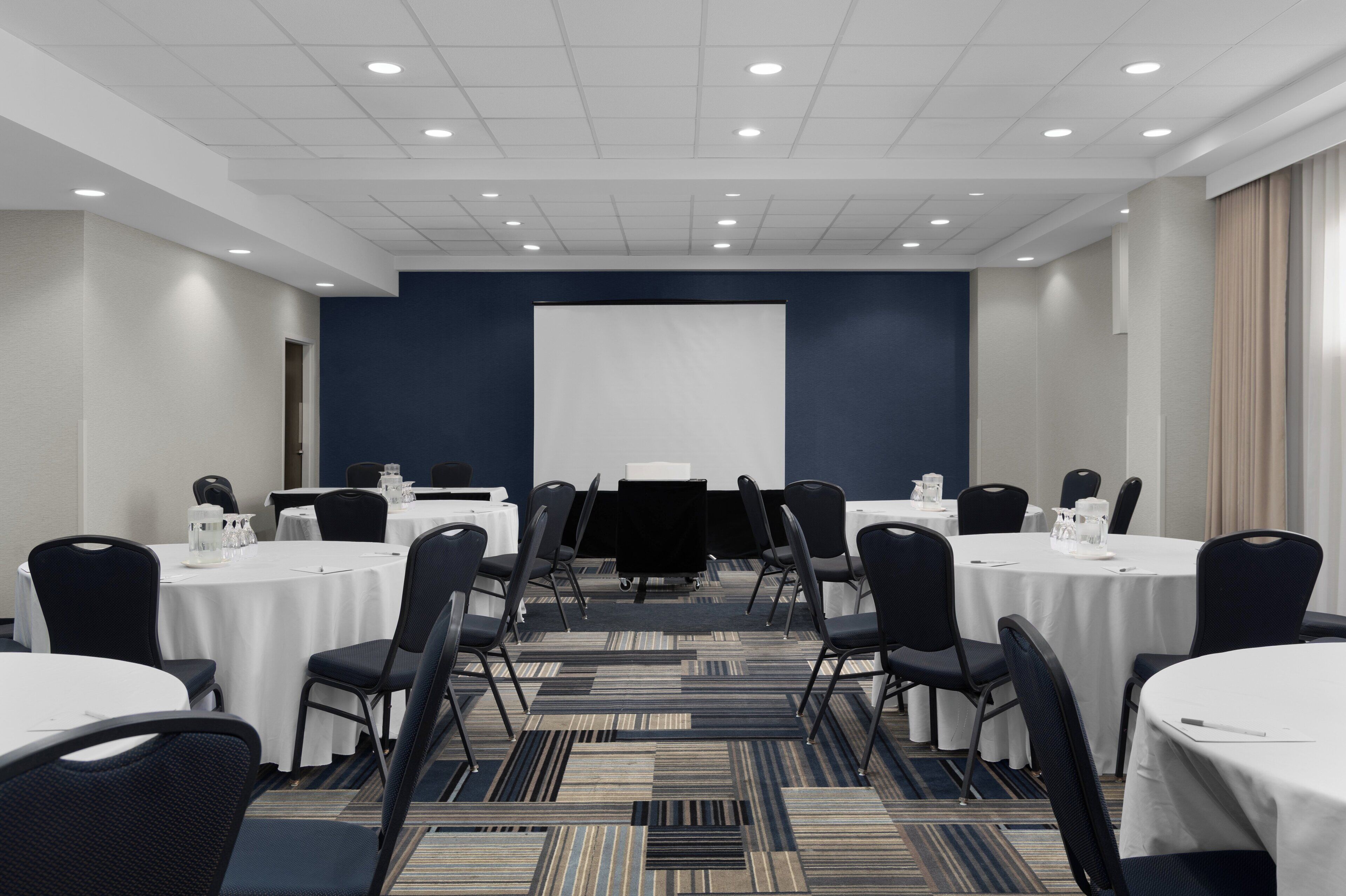 Une salle de conférence dot ée de plusieurs tables et chaises, d’un écran de projection et d’un sol recouvert de moquette.