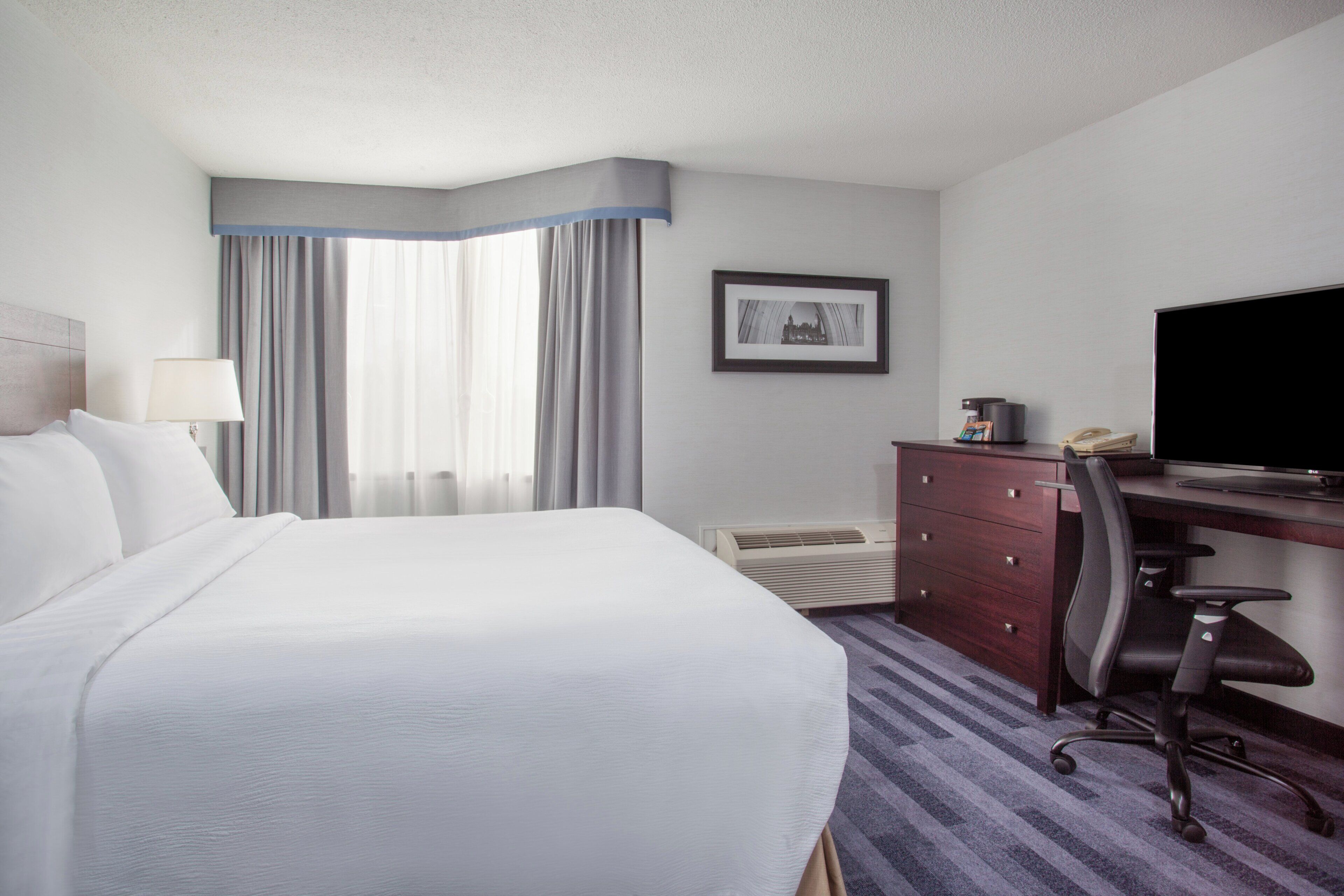 Habitación de hotel con una cama grande, un escritorio con computadora, una silla, unidad de aire acondicionado y un cuadro en la pared.