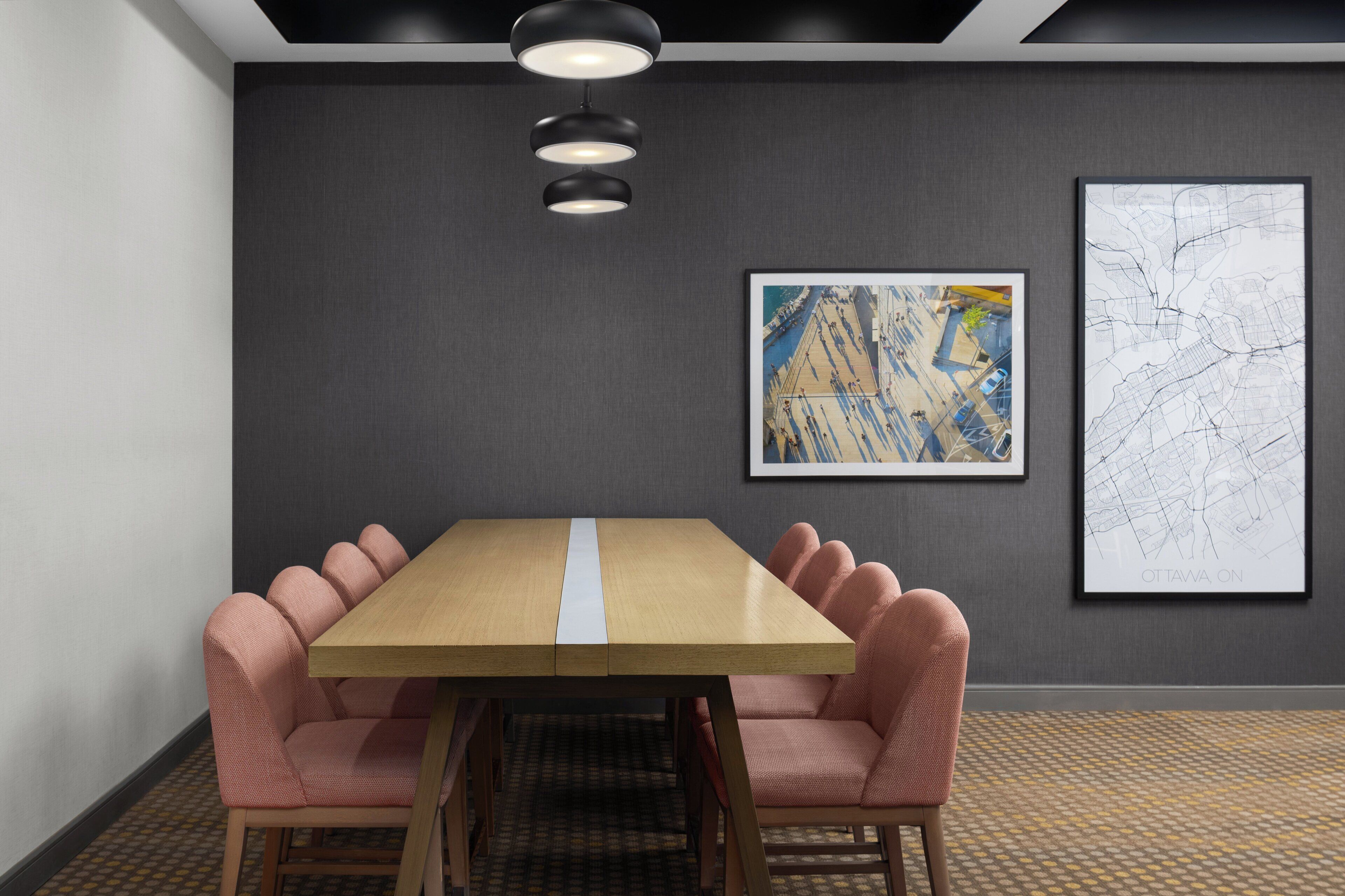 Uma sala de conferências com uma mesa de madeira, cadeiras estofadas em tons de rosa, um tapete estampado, uma luminária de teto preta e dois mapas emoldurados na parede.