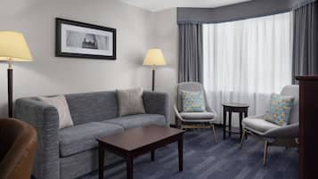 Une chambre d’hôtel avec un canapé gris, deux fauteuils, une petite table et une lampe. Un tableau encadré est accroché au mur, et il y a une fenêtre avec des rideaux.