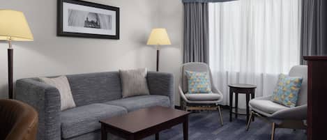 Hotelkamer met een grijze bank, twee fauteuils, een klein tafeltje en een lamp. Er hangt een ingelijst schilderij aan de muur en er is een raam met gordijnen.