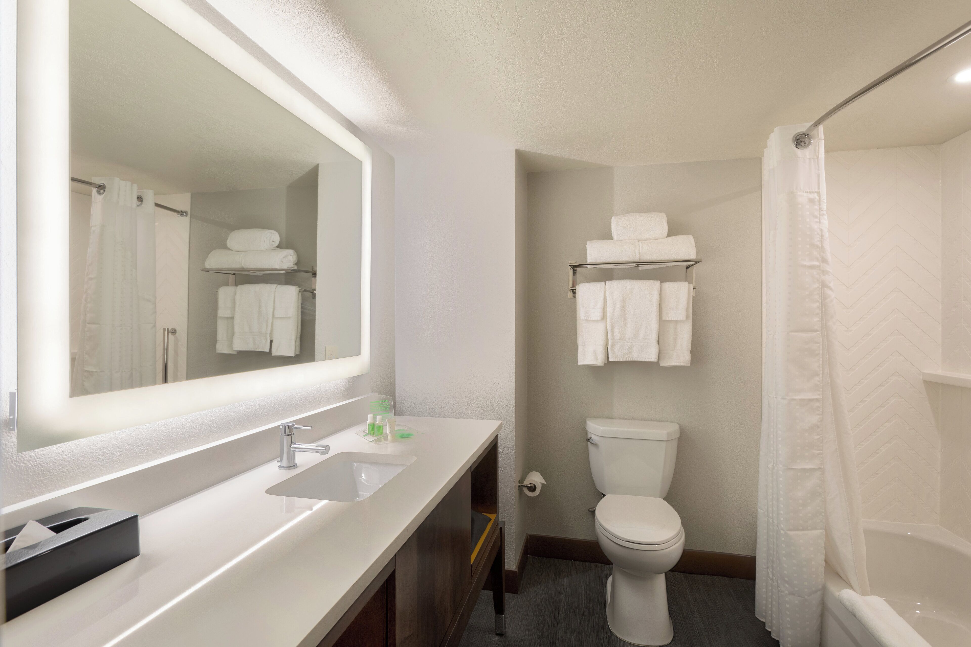 A modern bathroom with a white sink, mirror, and toilet. There are towels on a shelf above the toilet and a shower curtain.