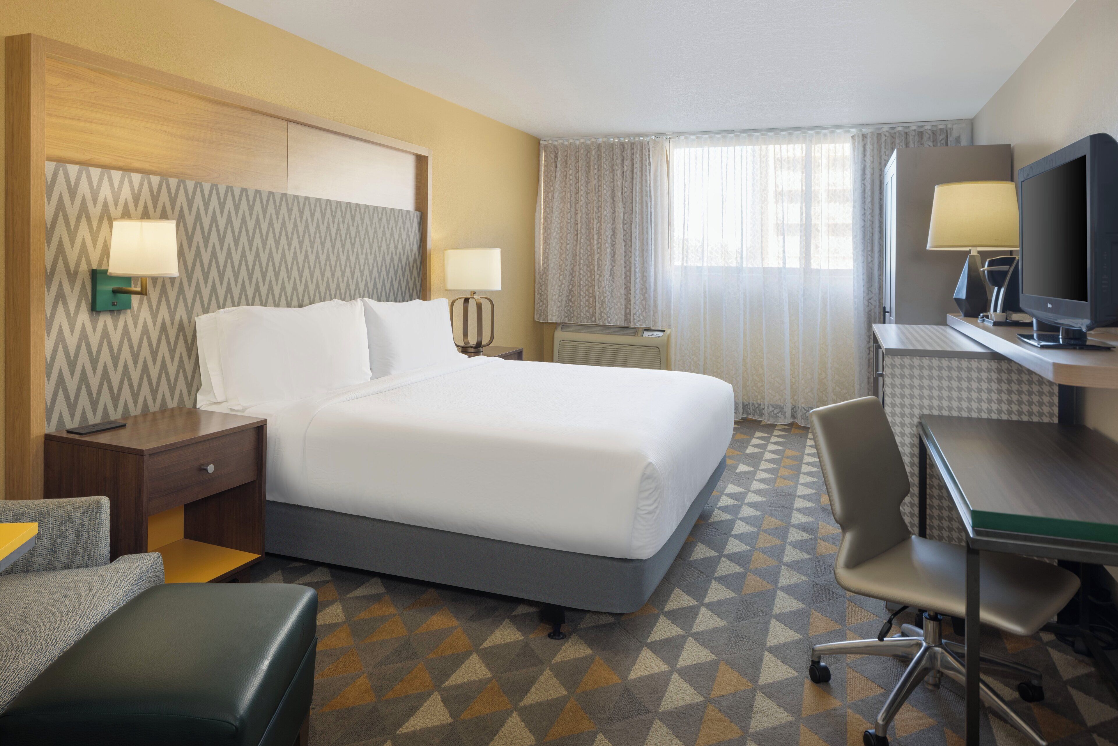 Habitación de hotel con cama, escritorio con computadora, silla, televisor y ventana con cortinas.