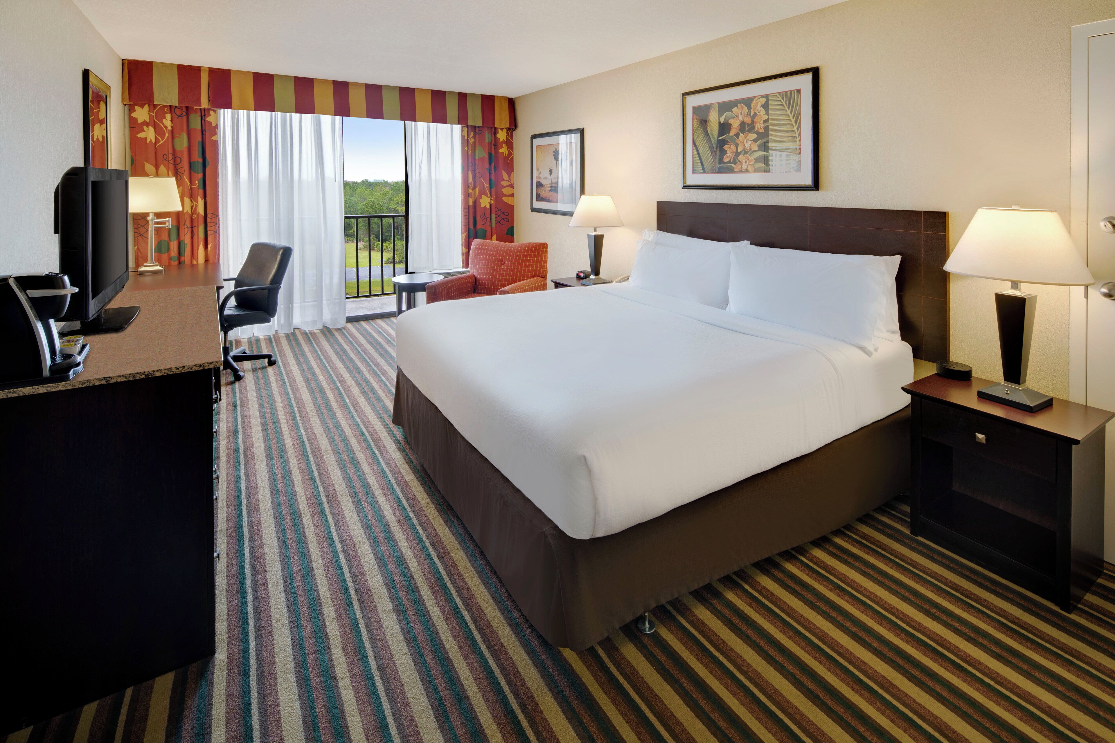 Une chambre d’hôtel avec un grand lit, un bureau avec une télévision, deux chaises, une fenêtre avec des rideaux et un tableau au mur.