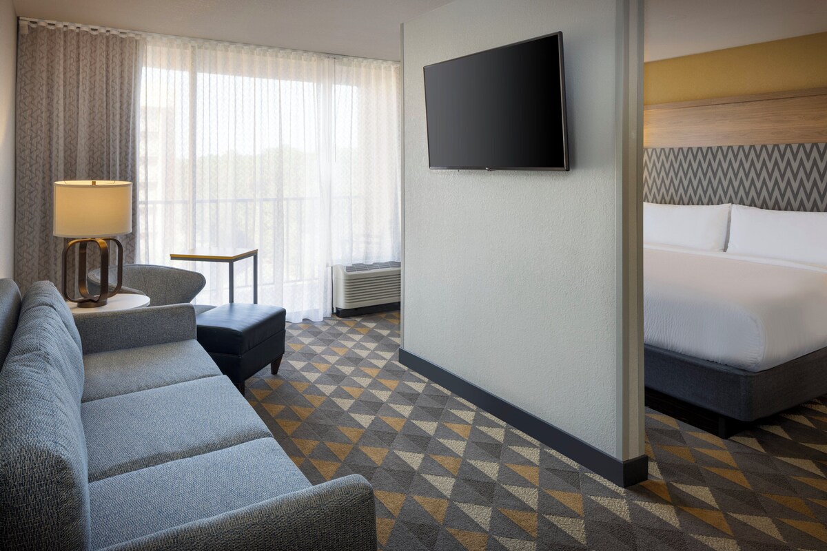 Quarto de hotel com sofá, televisão fixada na parede, cama com cabeceira e carpete estampado.