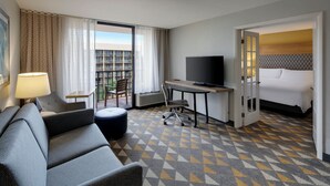 Une chambre d’hôtel avec un canapé, un bureau avec une télévision, un lit et une vue sur un immeuble doté de balcons.