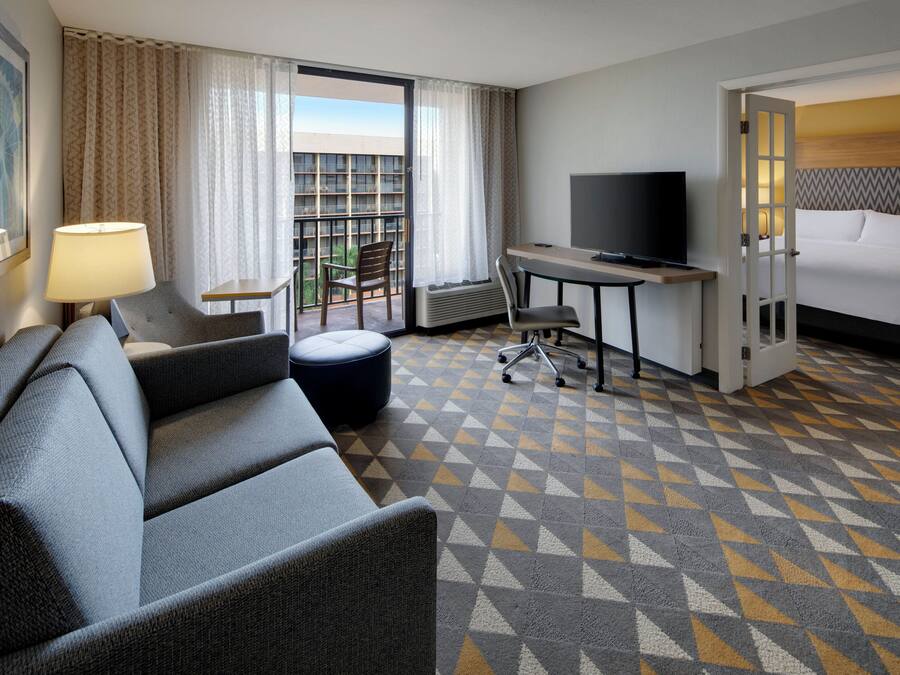 A hotel room with a sofa, a desk with a TV, a bed, and a view of a building with balconies.