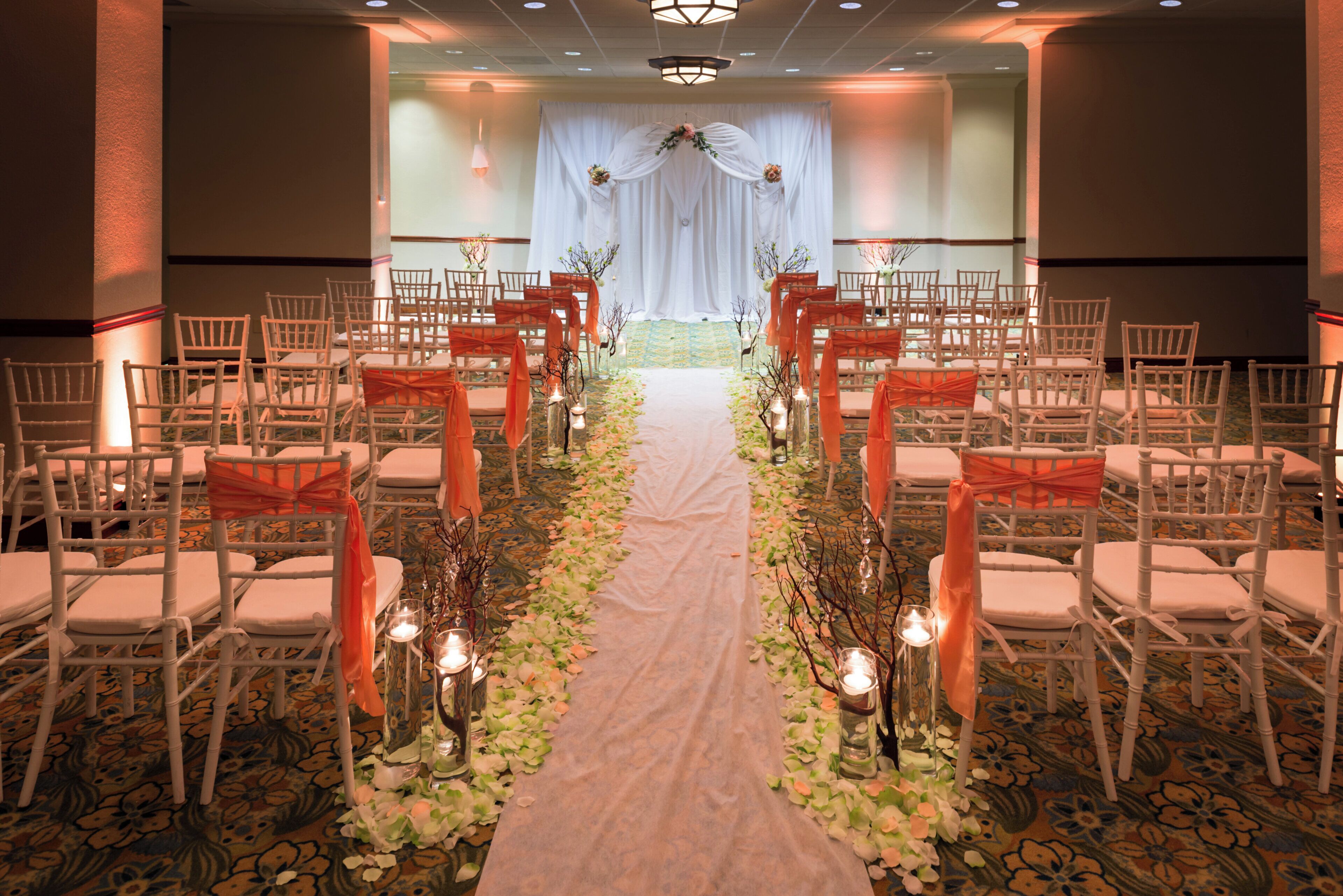 Ein Hochzeitgang mit Stühlen an beiden Seiten, einem weißen Vorhang im Hintergrund und Blumenschmuck.