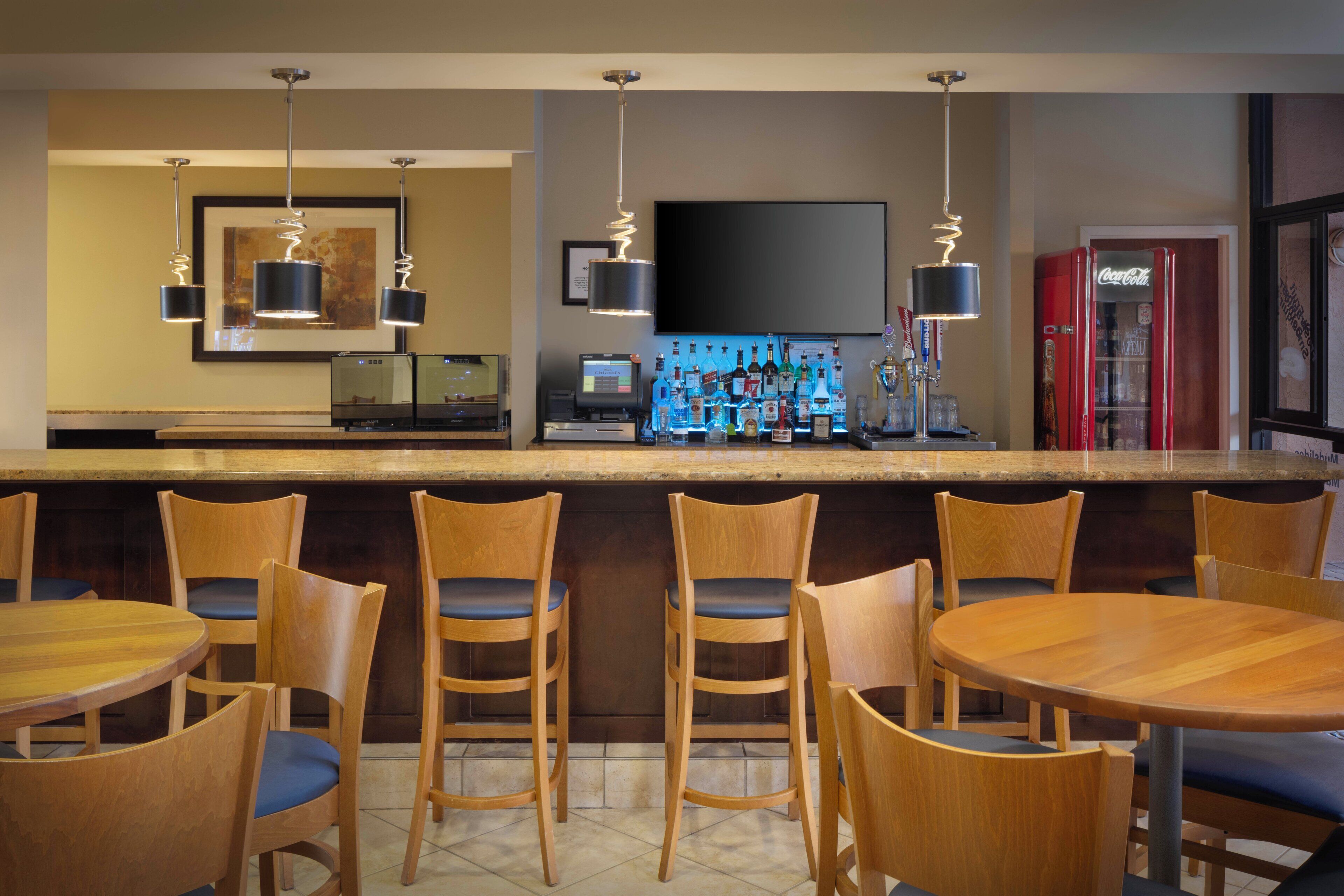 A bar area with wooden chairs and tables, a bar counter with various bottles, and a television mounted on the wall.