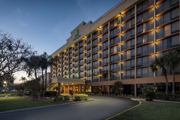 Um hotel grande com vários andares, entrada bem iluminada e entrada por um jardim.