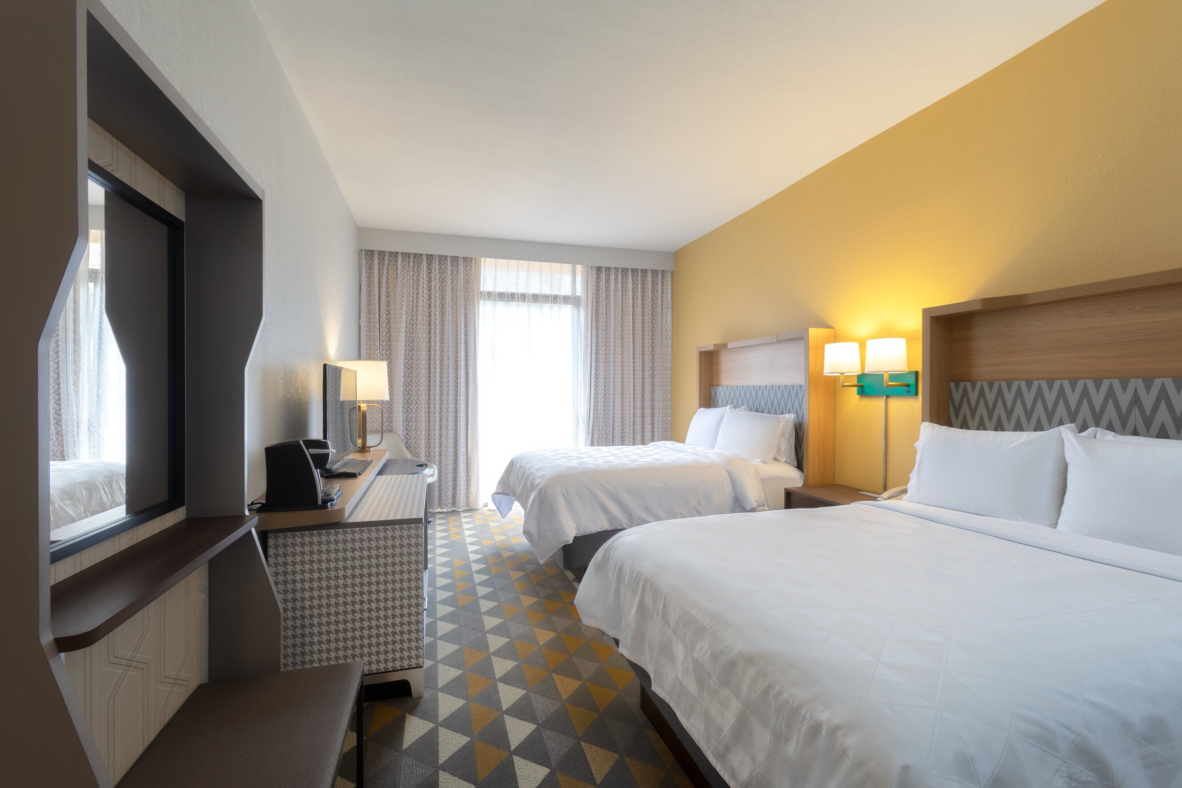 A hotel room with two beds, a desk, and a chair. There is a window with curtains, a lamp, and a wall-mounted light fixture.