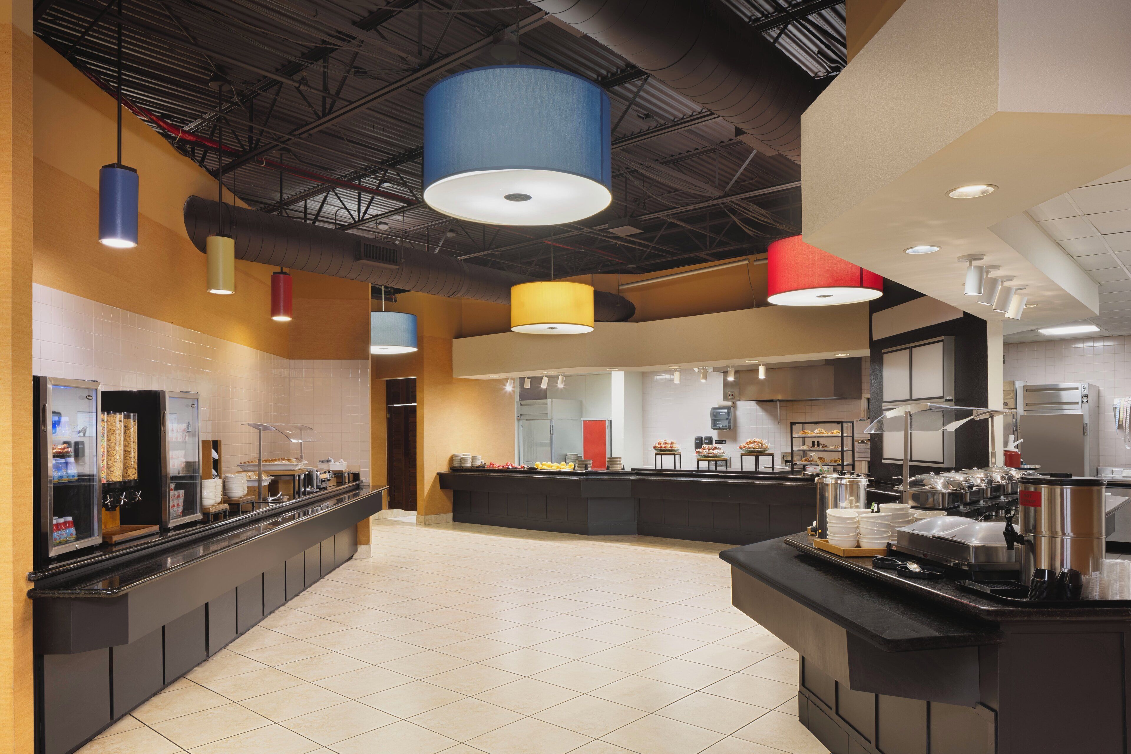 A modern hotel lobby with a long reception counter, a central island with food and drink stations, and a high ceiling with exposed ductwork.
