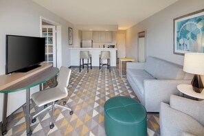 Quarto de hotel moderno com televisão de tela plana, escrivaninha, sofá confortável, mesinha e área de cozinha com um banquinho.