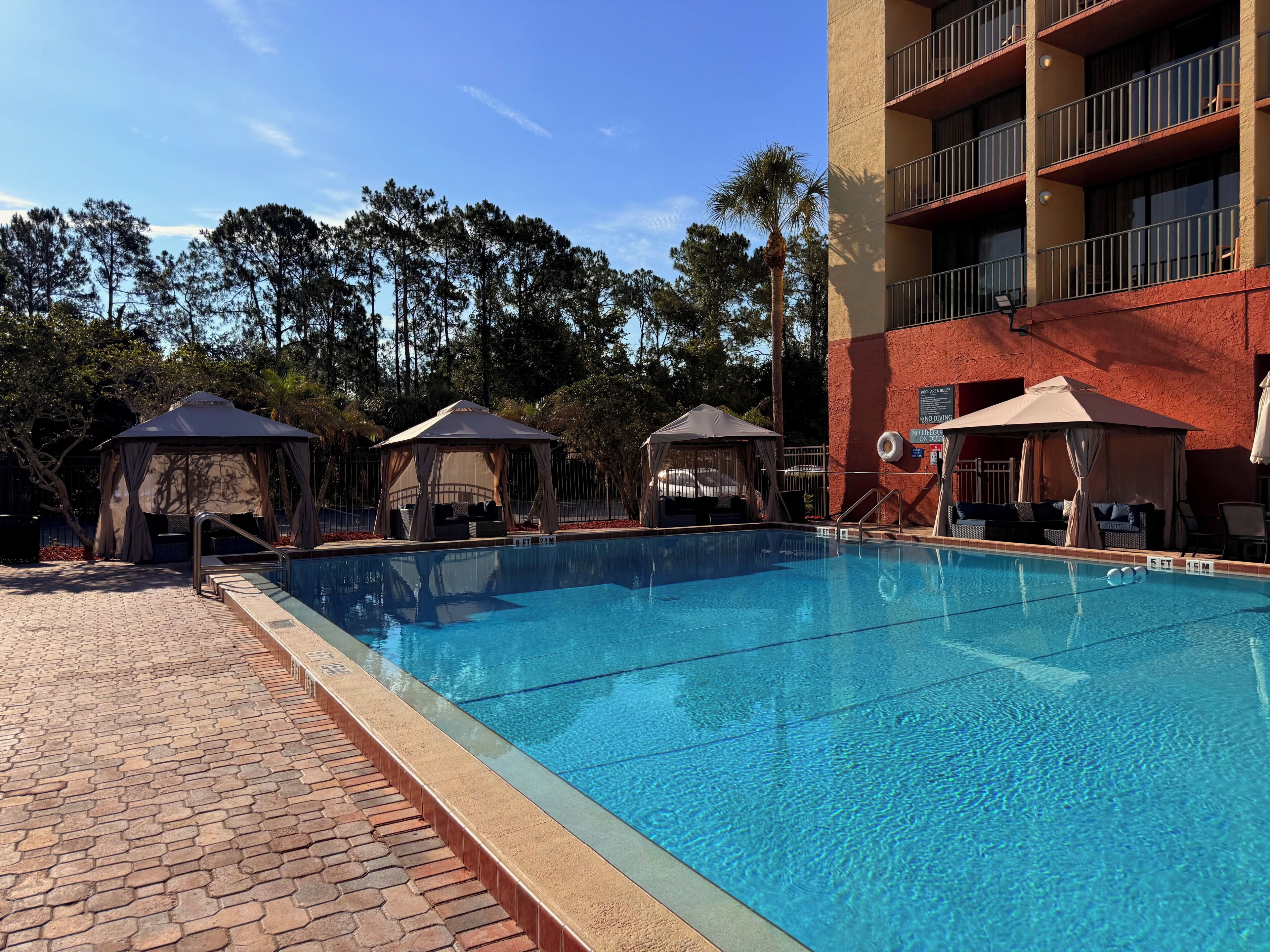 A swimming pool with lounge chairs and gazebos, surrounded by trees and a building with balconies.