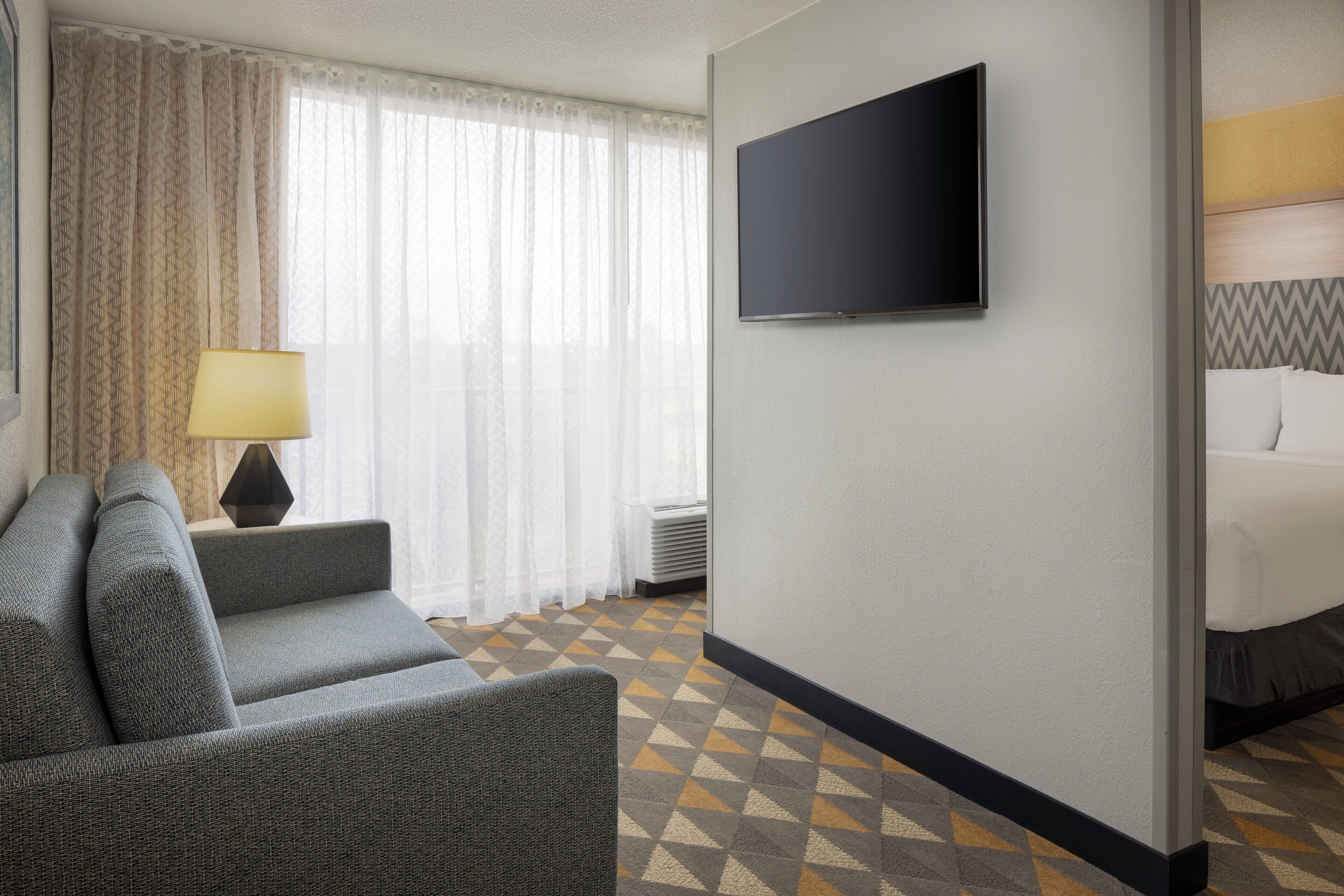 Hotellihuone, jossa on sohva, seinälle asennettu televisio, päätyä sisältävä sänky ja kuviollinen matto.