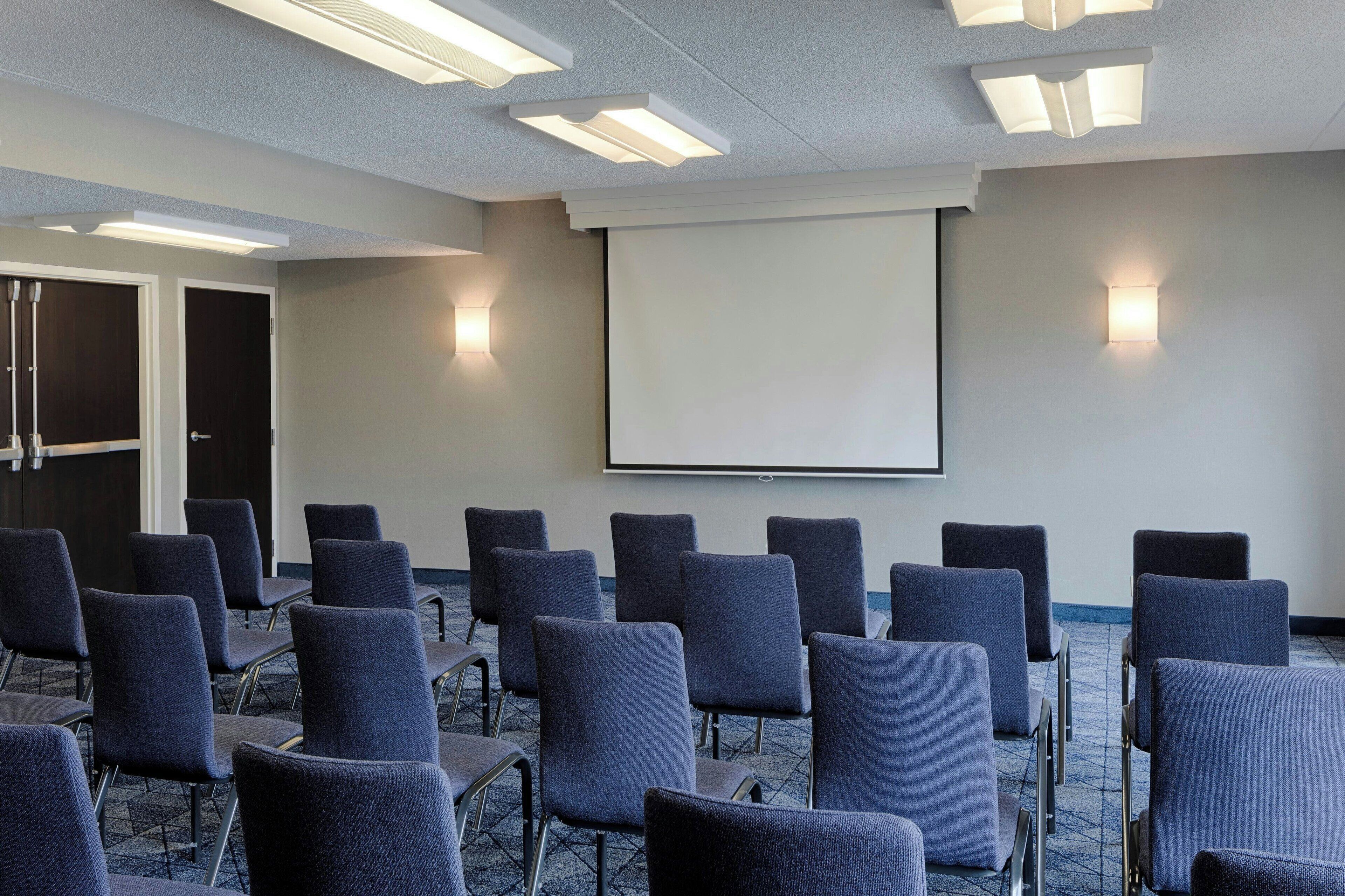 A conference room with rows of chairs, a projector screen, and wall-mounted lights.