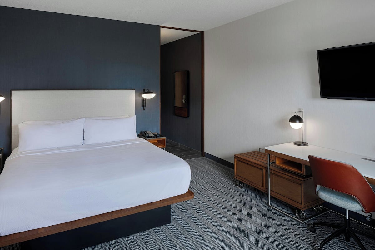 A hotel room with a large bed, a desk with a chair, a flat-screen TV, and a nightstand with a lamp.