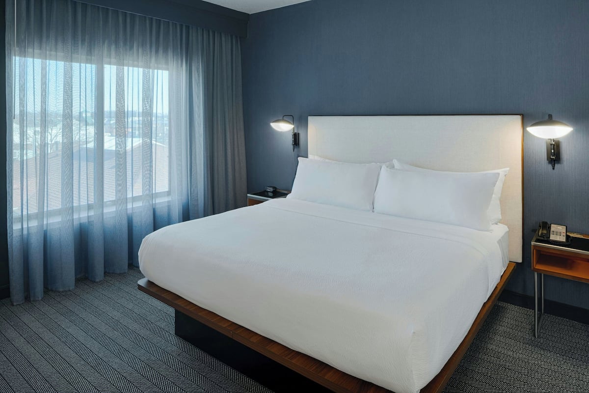 A hotel room with a large bed, a nightstand, a lamp, and a view through sheer curtains.