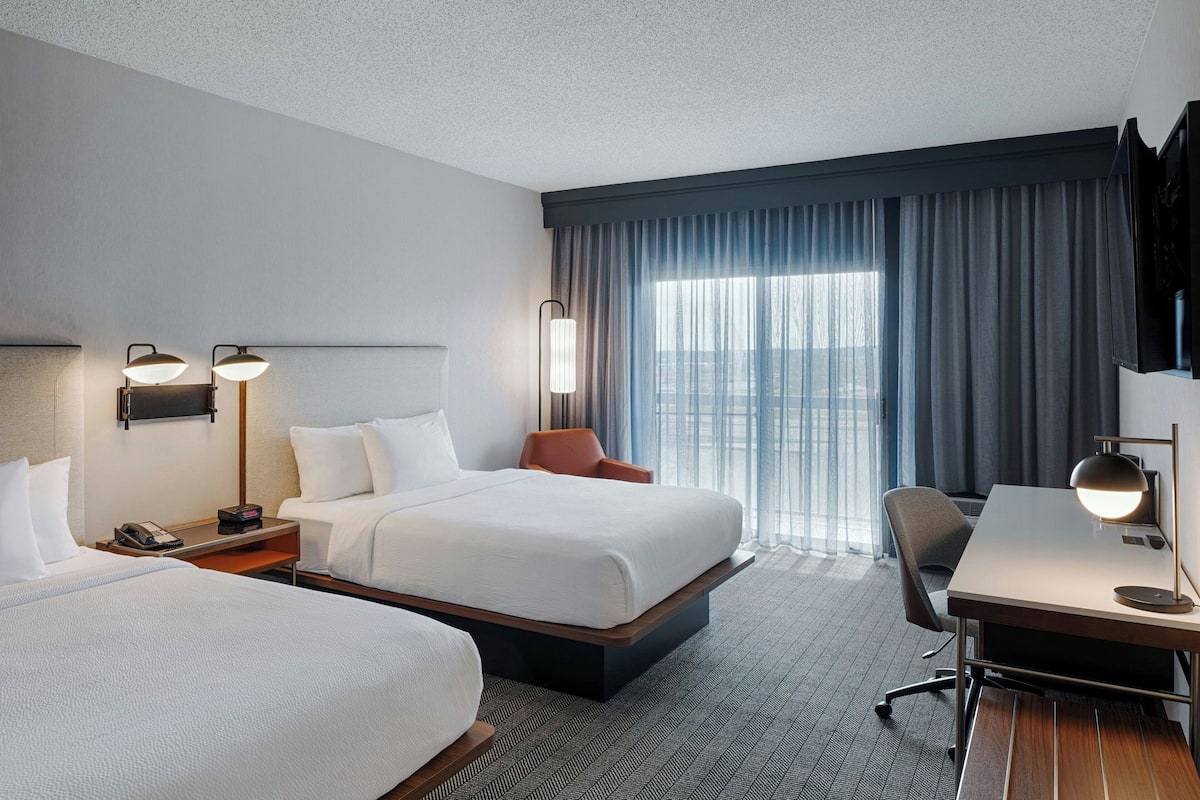 A hotel room with two beds, a desk, a chair, and a large window with curtains.