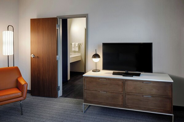 Une chambre d’hôtel avec une porte en bois, un téléviseur posé sur une console blanche, une lampe moderne et un tapis à motifs.