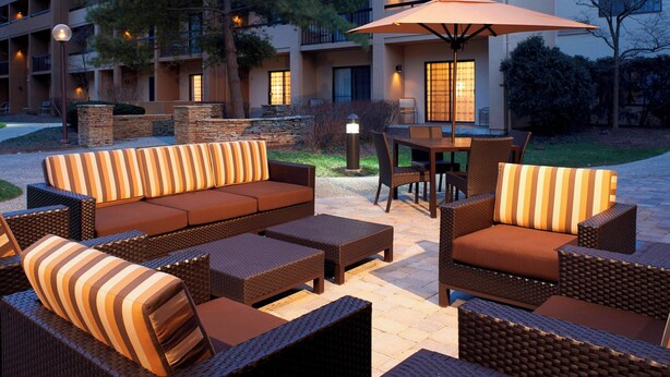 An outdoor seating area with wicker furniture and a table under an umbrella, surrounded by a building with multiple balconies and lights.