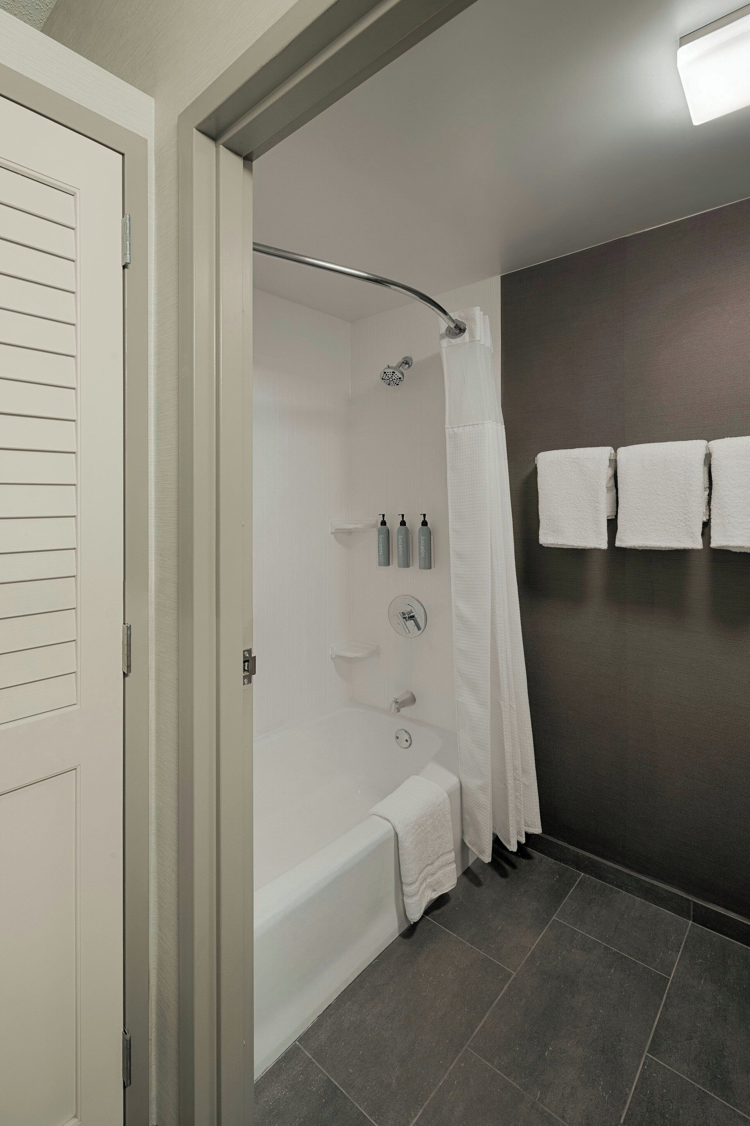 A modern bathroom with a bathtub, shower, and toilet. There are white towels hanging on the wall and toiletries on the wall-mounted shelves.