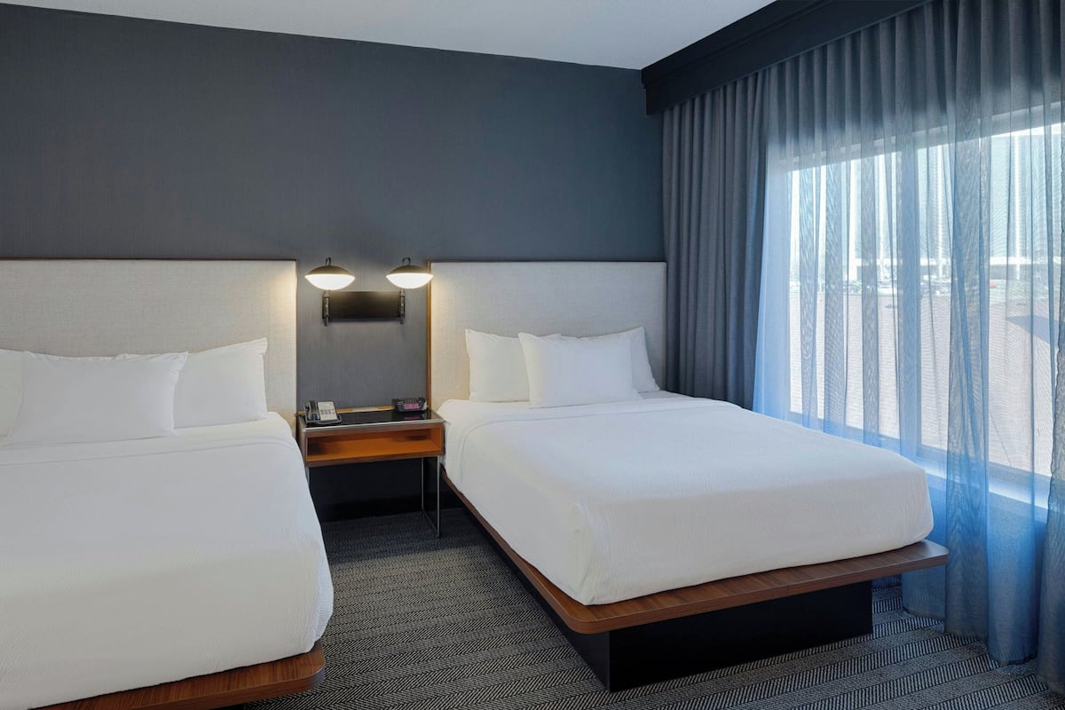 A hotel room with two beds, a nightstand with a lamp, and a large window with curtains.