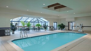 An indoor swimming pool with clear glass walls, surrounded by potted plants and outdoor seating.