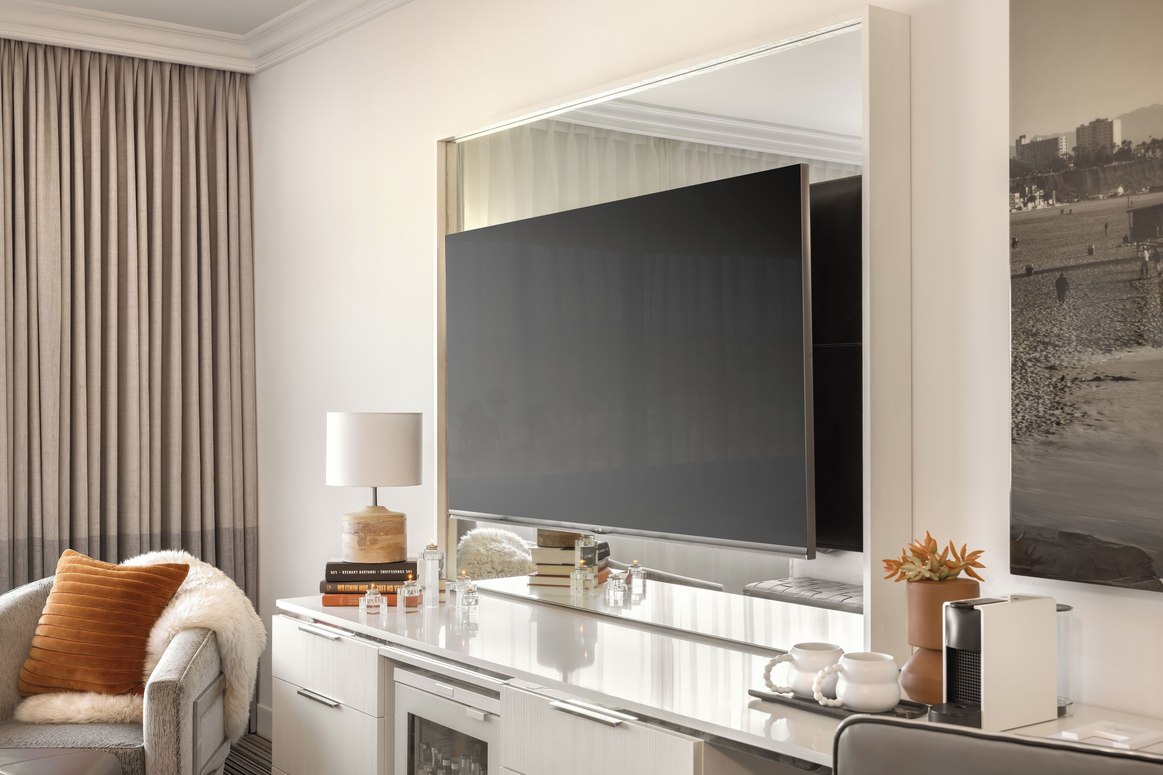 A modern hotel room with a large mirror, a desk with a lamp, a chair, and a potted plant.