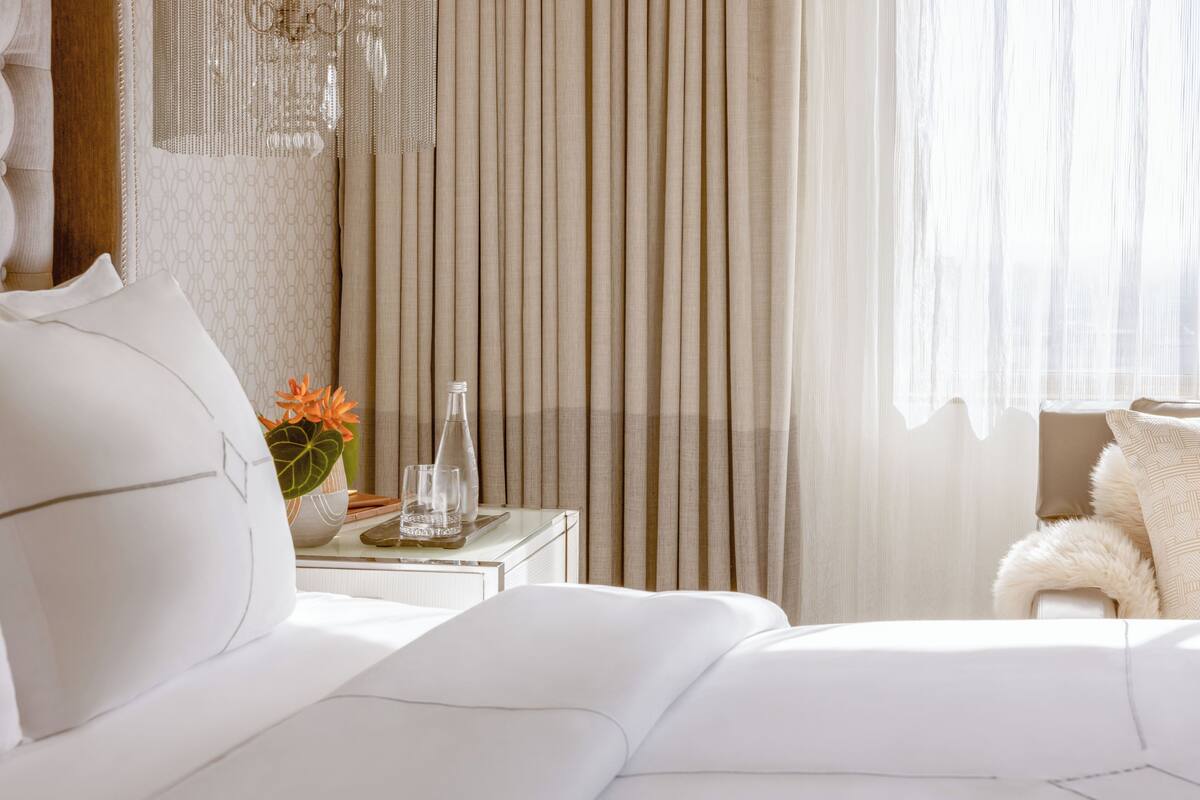 A hotel room with a neatly made bed, a bedside table with a vase of flowers, a glass of water, and a lamp.