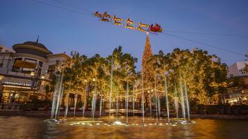 A fountain with water jets, surrounded by trees decorated with lights, and a building with a dome in the background.
