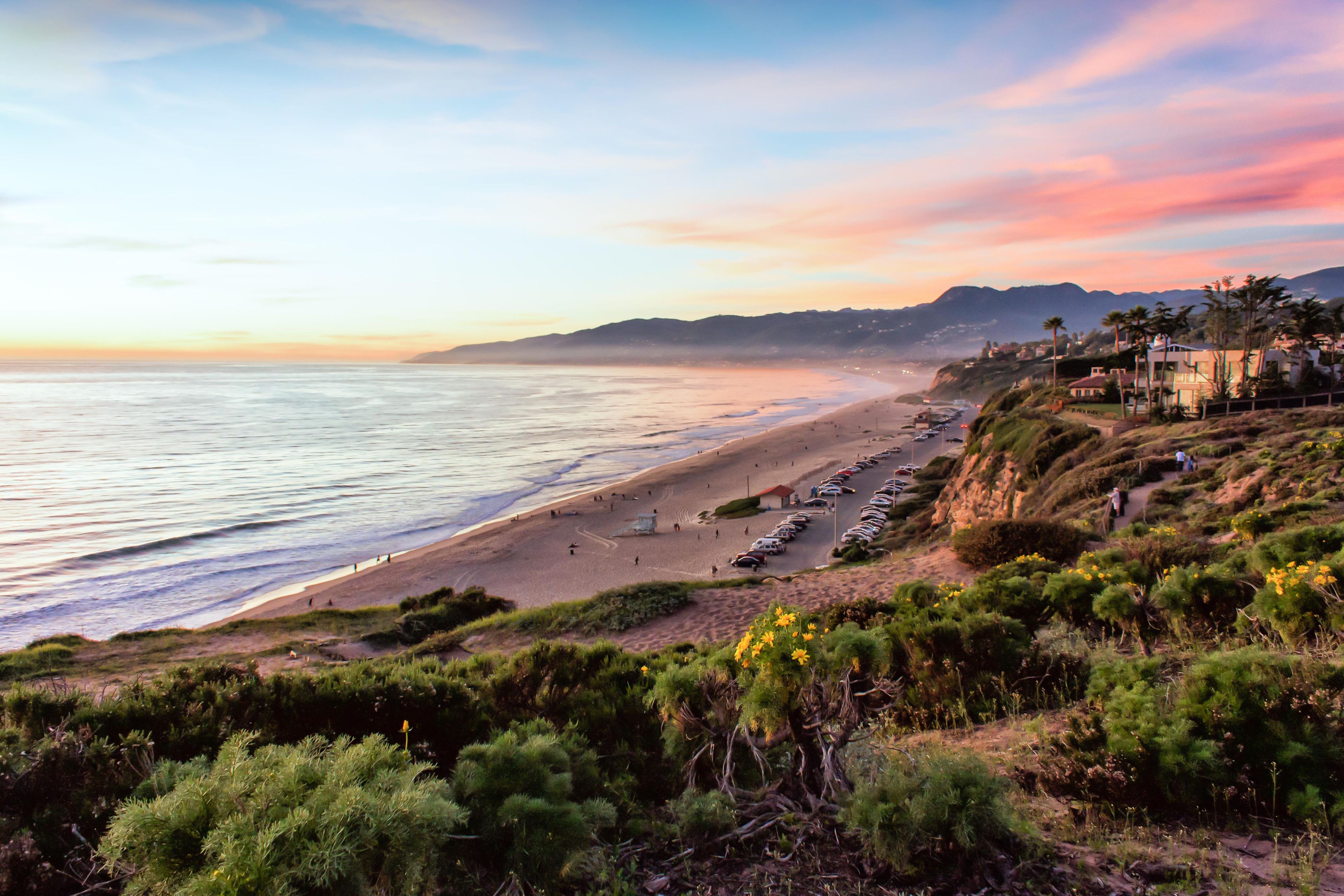 A coastal landscape with a beach, parked cars, and a hilly terrain with vegetation.