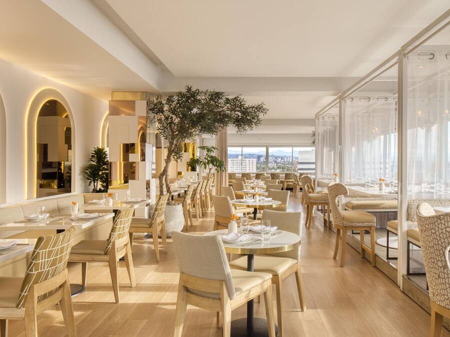 Um restaurante moderno com pé direito alto, grandes janelas e vista para a paisagem urbana.