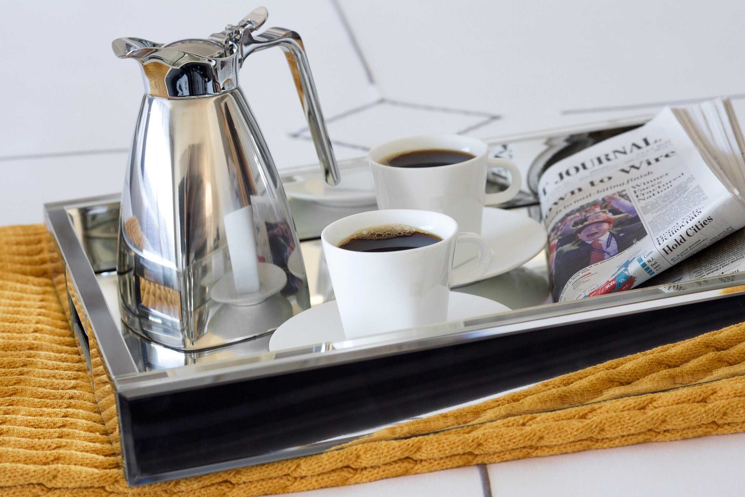 A tray with a silver coffee pot, two white cups of coffee, and a newspaper on a textured surface.