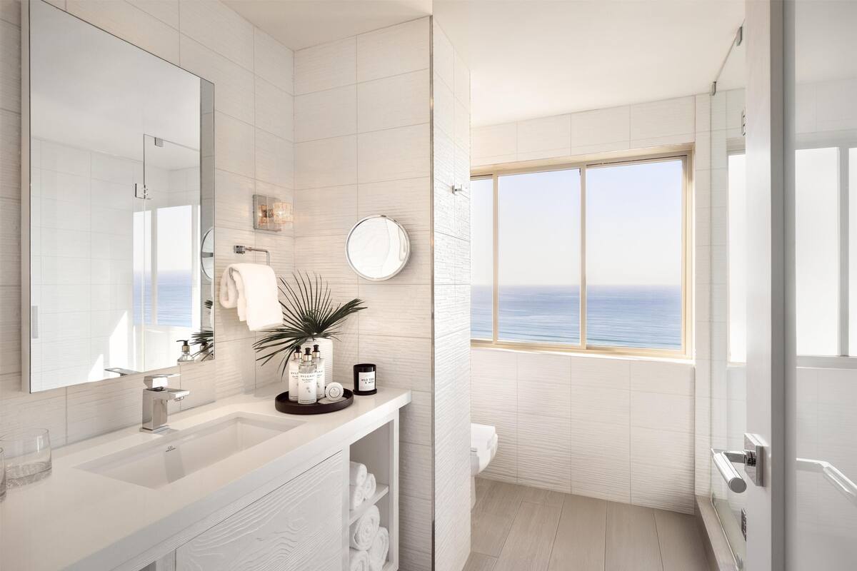 A modern bathroom with a large mirror, white tiles, a sink, and a window with a view of the sea.