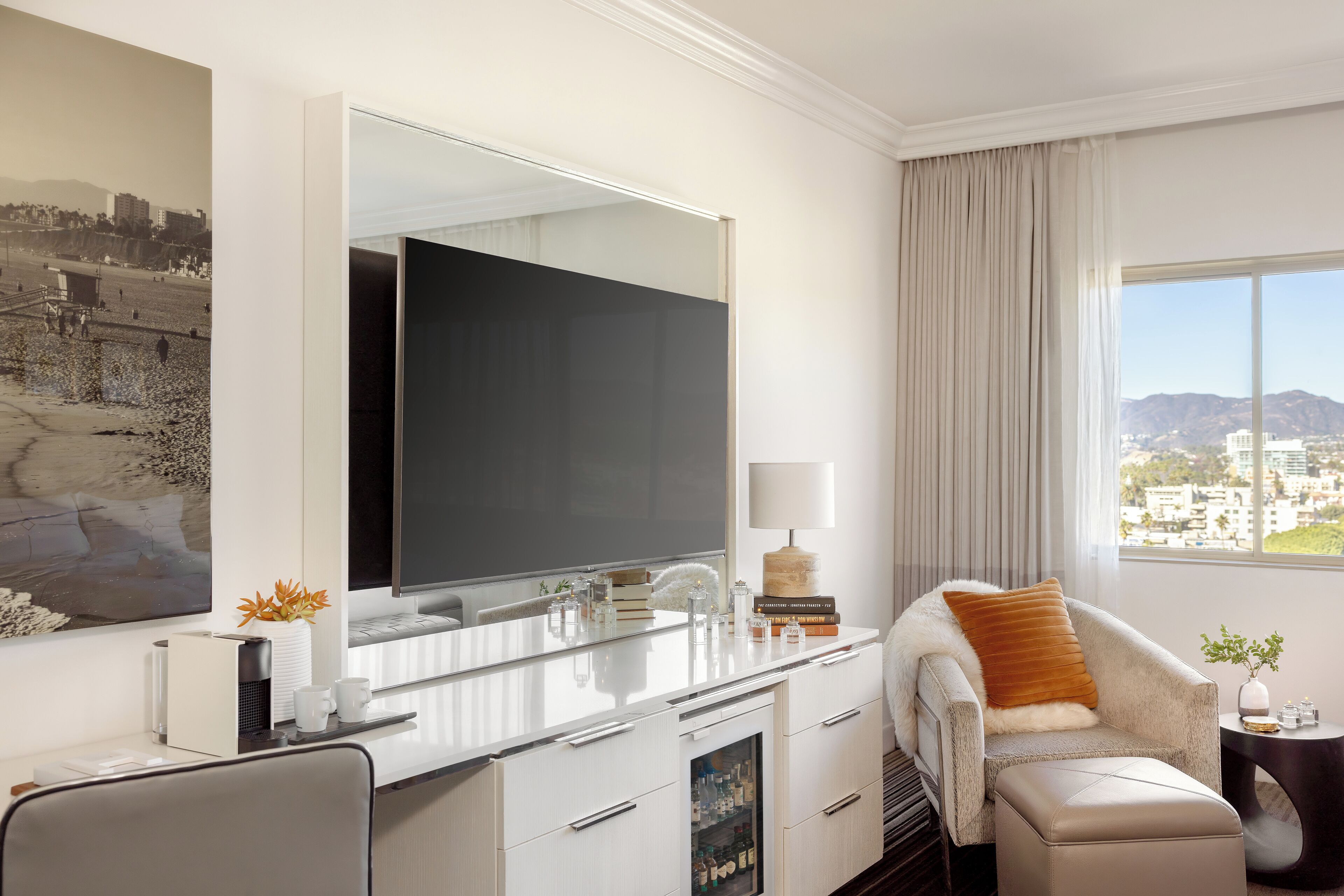 A modern hotel room with a large mirror, a flat-screen TV, a comfortable armchair, a small side table, and a view of the city.