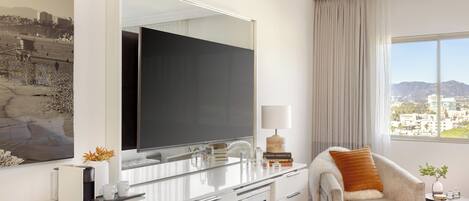 A modern hotel room with a large mirror, a flat-screen TV, a comfortable armchair, a small side table, and a view of the city.