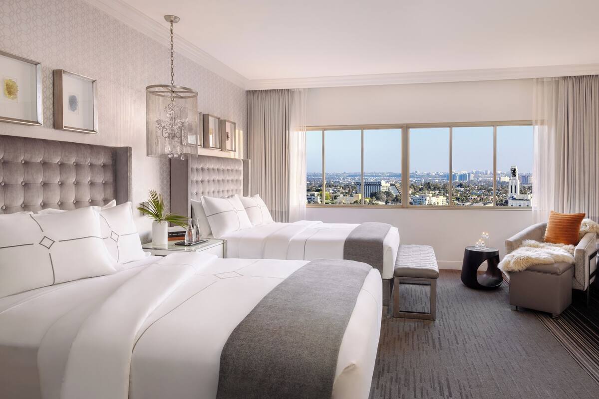 A hotel room with two beds, a large window with a city view, a chandelier, and a view of a tower.