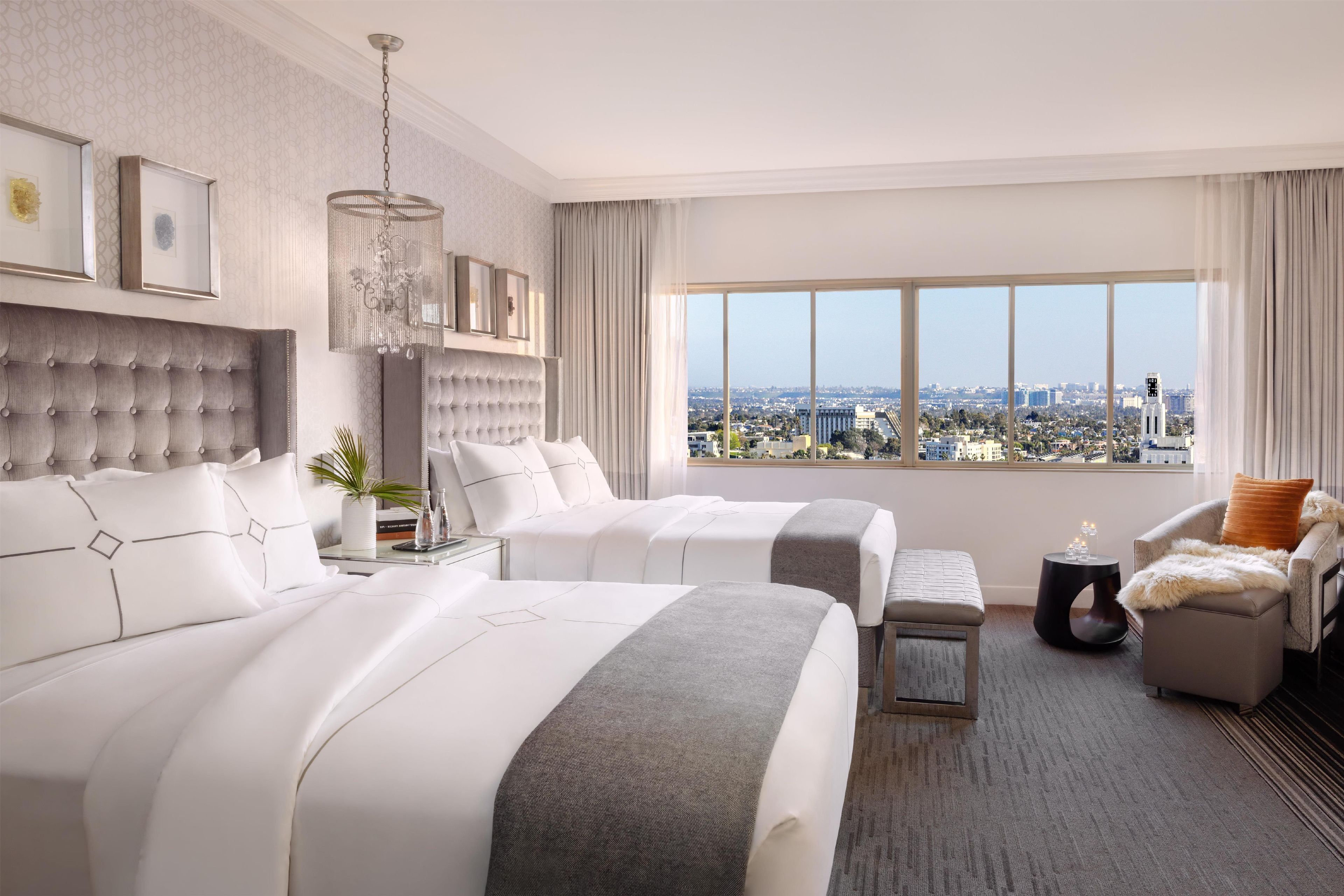 A hotel room with two beds, a sitting area, a view of the city, and a chandelier.