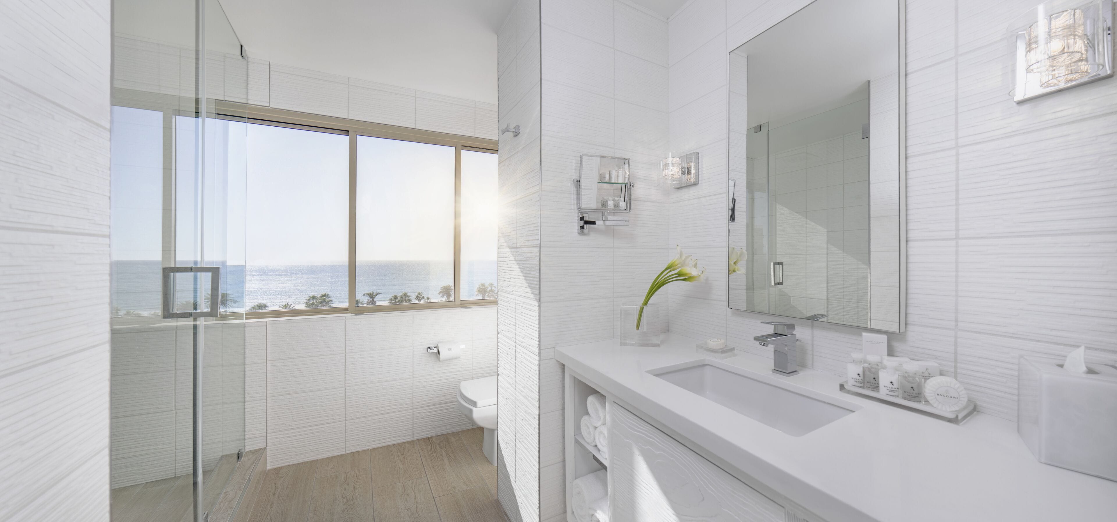 A modern bathroom with a large window, a glass shower enclosure, and a vanity with toiletries.