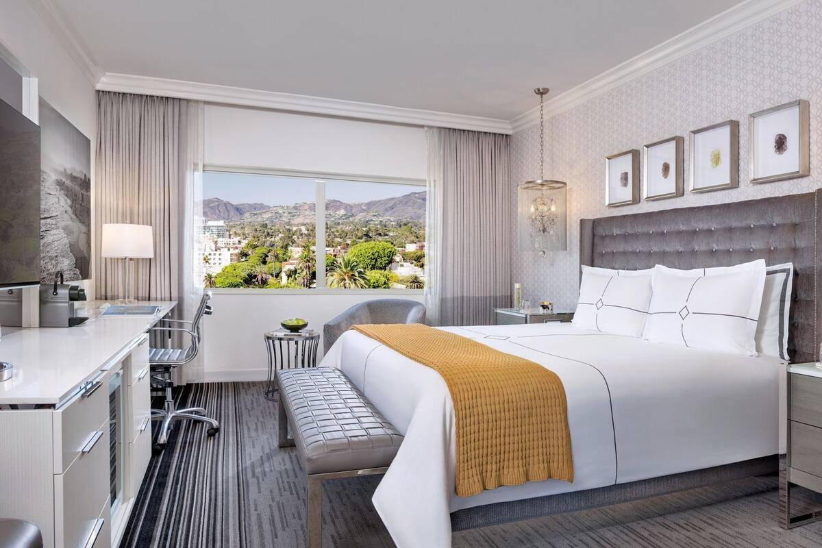 A hotel room with a bed, a desk, a chair, a bench, a view of the city, and a chandelier.