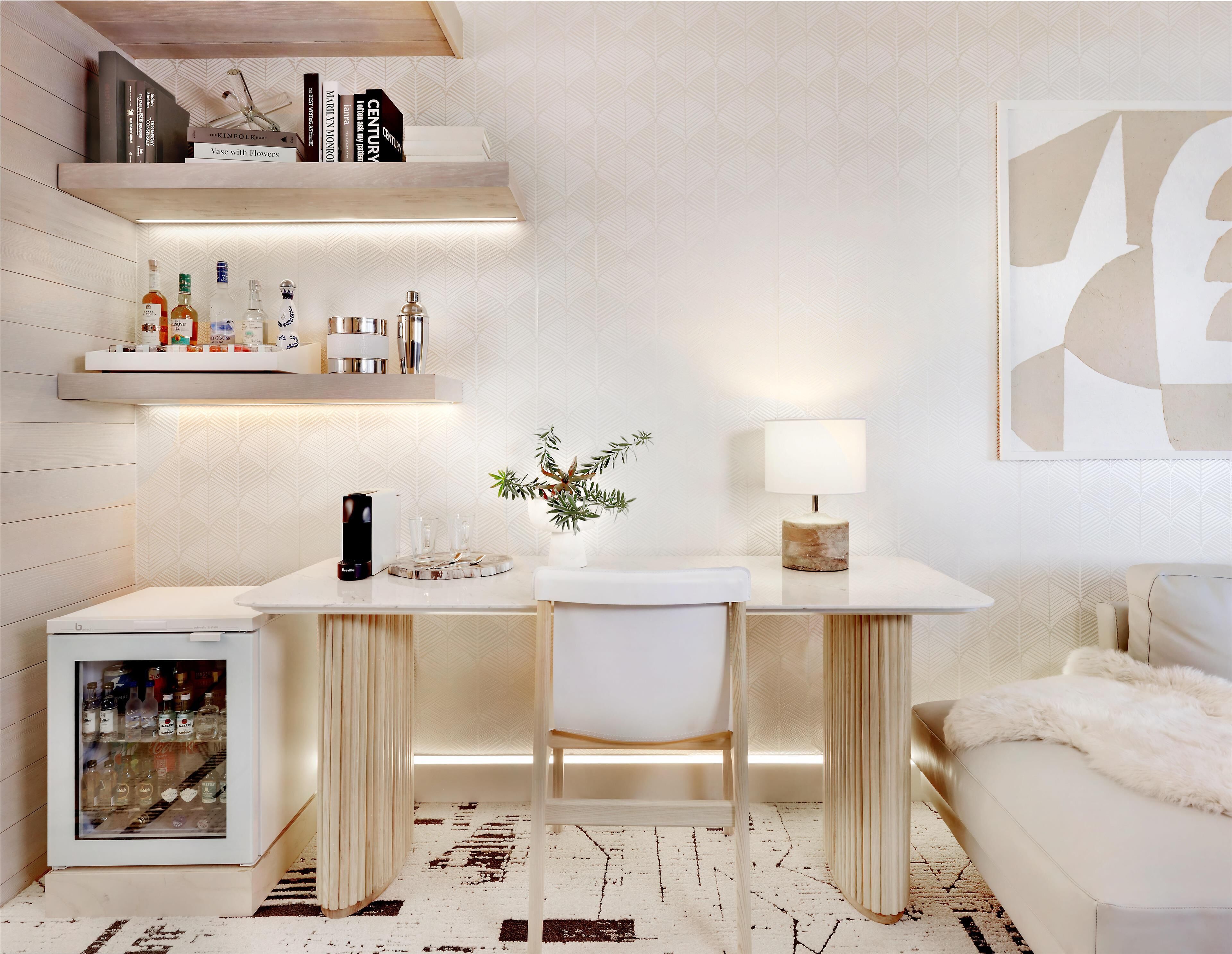 Un espace de travail moderne et bien éclairé, avec un bureau, une chaise et des étagères blancs. On aperçoit un petit réfrigérateur contenant des boissons, et une plante décorative apporte une touche de verdure.